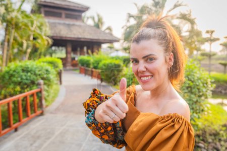 A white woman wears African clothing and gives a thumbs up while standing in a public park.