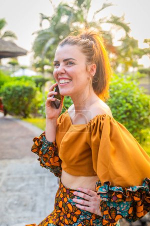 Una joven caucásica sonriente hace una llamada telefónica afuera, foto vertical.