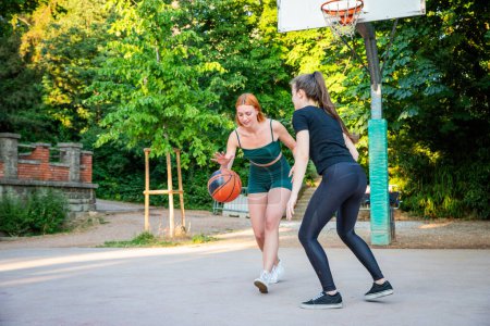 Two women playing basketball in a park. One of them is wearing a green shirt and shorts. The other is wearing a black shirt and black pants