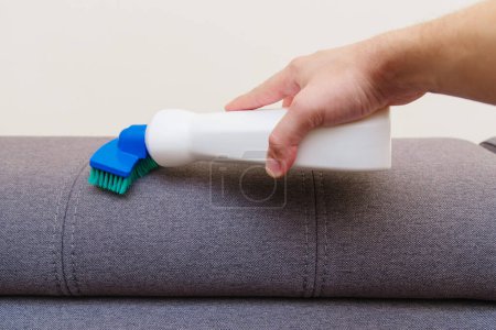 Foto de El hombre utiliza un producto de limpieza con una esponja y un cepillo para limpiar la superficie de un sofá. Limpiando la casa. Servicio de limpieza y limpieza. - Imagen libre de derechos