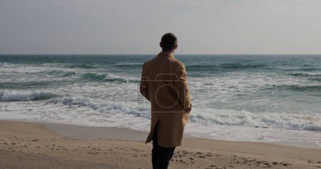 Téléchargez les photos : Homme méconnaissable en manteau regardant la mer debout sur le littoral, vue de dos - en image libre de droit