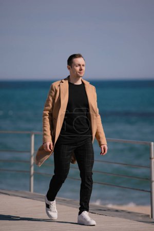 Foto de Hombre con estilo en el abrigo beige caminando a lo largo de la orilla del mar - Imagen libre de derechos