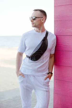 Téléchargez les photos : Bel homme de mode posant avec sac banane appuyé sur le mur en bois rose dans le costume blanc - en image libre de droit