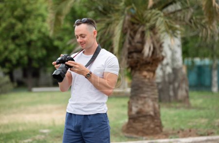 Photo for Man photographer looking photos in camera at the summer park with palm tree on background - Royalty Free Image