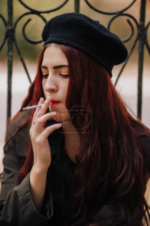 Photo for A girl in retro coat french style smoking cigarette sitting on fence in the autumn park - Royalty Free Image