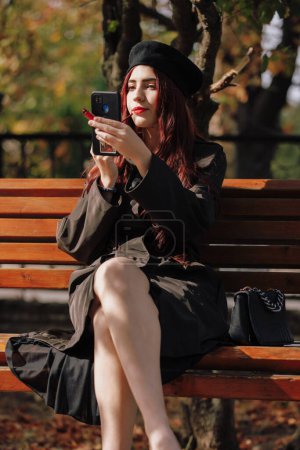 Téléchargez les photos : Une fille en manteau s'assoit sur un banc et peint ses lèvres dans un parc d'automne - en image libre de droit
