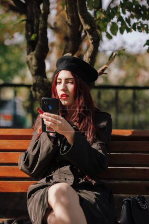Téléchargez les photos : Une fille en manteau s'assoit sur un banc et peint ses lèvres dans un parc d'automne - en image libre de droit
