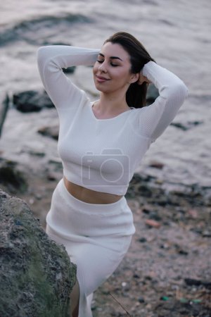 Téléchargez les photos : Élégante dame en robe blanche posant sur des pierres près de la mer - en image libre de droit