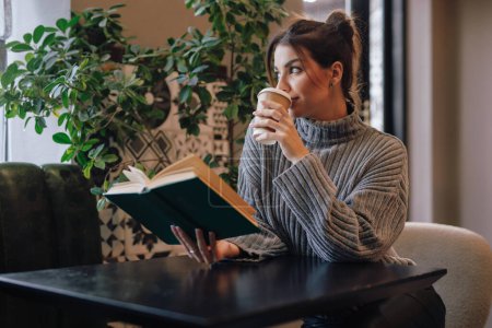 Photo for Young woman reading book and drinking coffee sitting in a cafe at the table - Royalty Free Image