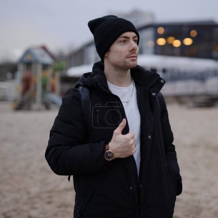 Téléchargez les photos : Jeune homme de mode en veste d'hiver noire et chapeau sur la plage - en image libre de droit
