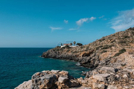 Foto de Paredes encaladas y techo de baldosas de la Iglesia de Sotiros cerca de Loutro, Creta se destacan en la costa escarpada y rocosa contra el cielo azul y el mar. - Imagen libre de derechos