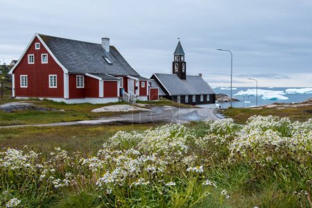 Téléchargez les photos : Des fleurs sauvages occupent le premier plan de cette vue sur l'église de Sion à Illulisat, au Groenland. En arrière-plan se trouvent des icebergs du massif Icefjord. - en image libre de droit