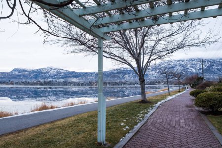Paved footpath along Osoyoos Lake waterfront in winter.  Perfectly calm lake water produces mirror-like reflection of opposite shore and snow-covered mountains.