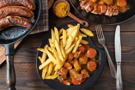 Traditional german fast food meal with currywurst or curry sausage with french fries and delicious curry sauce. Served on plates with cutlery on wooden and rustic table background. Flat lay