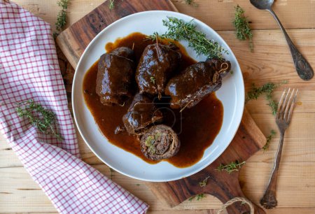 Rulades de carne alemana rellenas de encurtidos, tocino, cebolla y mostaza con deliciosa salsa marrón en bandeja sobre mesa de madera con vista transversal.