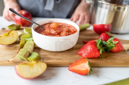 Fresh cooked rhubarb compote with strawberries and apples in a bowl on a cutting board with ingredients in the kitchen