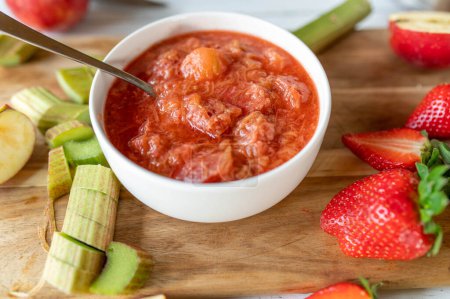 Frisch gekochtes Rhabarberkompott mit Erdbeeren und Äpfeln in einer Schüssel auf einem Schneidebrett mit Zutaten aus der Küche