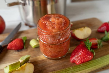 Frisch gekochtes Rhabarberkompott mit Äpfeln und Erdbeeren im Glas auf einem Holzschneidebrett in der Küche.