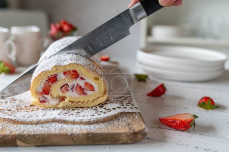 Frisch gebackenes Kuchenbrötchen oder Kuchenroulade wird von Frauenhand mit einem Küchenmesser geschnitten