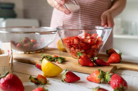 Woman is sweetening chopped strawberries with sugar in a glass bowl in the kitchen.