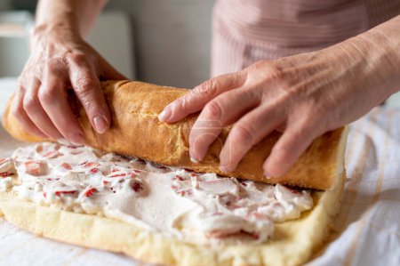 Zubereitung einer Tortenroulade oder einer Schweizer Rolle. In der Küche wird eine Torte mit Füllung von Frauenhänden aufgerollt. Frontansicht mit selektivem Fokus auf verschwommenem Hintergrund.