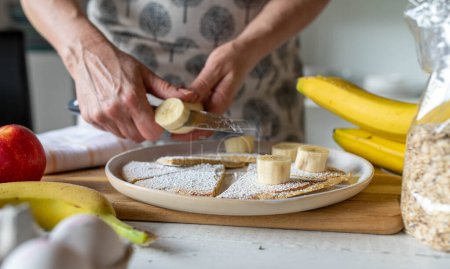 Womans hand  topping a fresh oatmeal pancake with bananas. Cutting bananas.