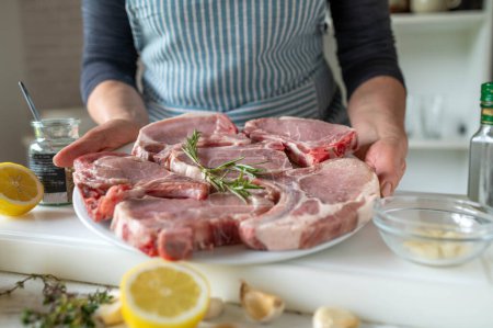 Chuletas de cerdo frescas y crudas listas para ser marinadas por una mujer con delantal en la cocina