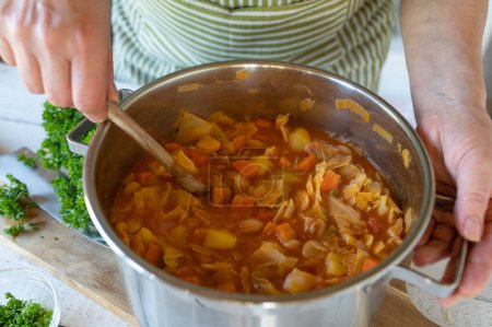 Healthy vegan soup is stirred with wooden spoon by womans hand int he kitchen. Closeup