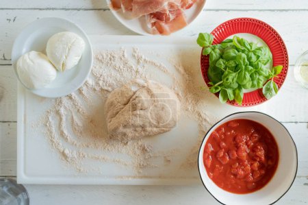 Vollkorn-Pizzateig auf bemehlter Oberfläche mit Zutaten wie Schinken, Basilikum, Mozzarella für die Zubereitung einer Pizza