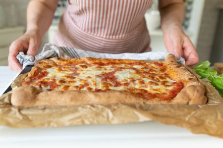 Woman with apron holding a fresh baked sheet pan pizza margherita in her hands on kitchen counter. Baked with whole wheat flour
