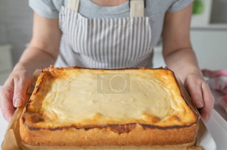 Femme avec tablier dans la cuisine tenant un gâteau au fromage frais en forme de carré dans ses mains.