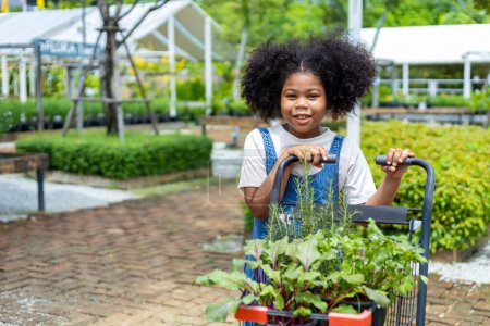 Téléchargez les photos : Portrait de l'enfant africain choisit des légumes et des plantes herbacées de la pépinière du centre de jardin local avec panier plein de plantes d'été pour le jardinage week-end et concept extérieur - en image libre de droit