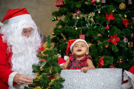Téléchargez les photos : Le bébé afro-américain s'amuse à rire et à jouer pendant que le Père Noël prépare un cadeau et est assis derrière l'arbre de Noël pour la célébration de la saison - en image libre de droit