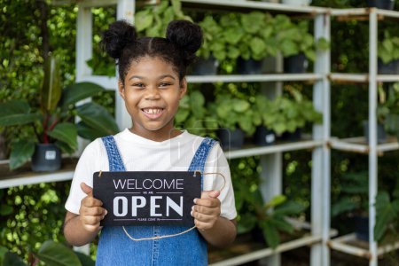 Foto de Retrato de niña afroamericana sosteniendo signo abierto de bienvenida para su negocio de centro de jardinería familiar para vivero y concepto de tienda de plantas de interior - Imagen libre de derechos