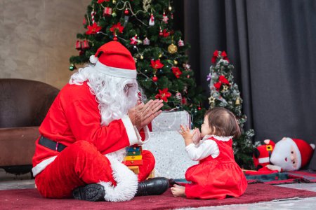 Photo for Toddler baby girl is having fun playing with Santa claus in the room fully decorated and christmas tree for season celebration - Royalty Free Image