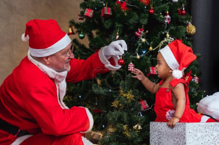 Photo for African American baby is getting ornamental bauble as present from Santa claus at night by the fully decorated christmas tree for season celebration concept - Royalty Free Image
