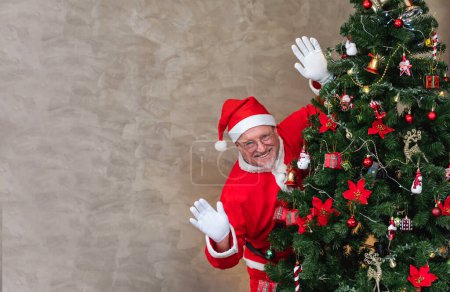 Foto de Santa Claus está celebrando la Navidad en la felicidad y la emoción mientras se esconde detrás del árbol de Navidad totalmente decorado y saludando para el saludo de temporada y la felicidad - Imagen libre de derechos