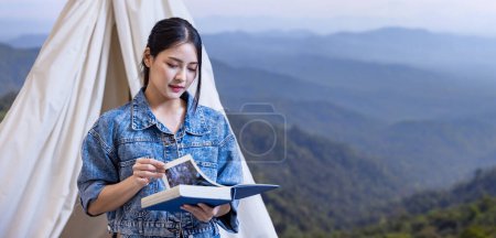 Foto de Asian woman reading book while on a solo trekking camp on the top of the mountain with small tent for weekend activities and outdoor pursuit concept - Imagen libre de derechos