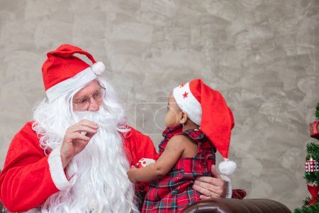 Téléchargez les photos : Père Noël soulève heureuse petite fille tout-petit et assis sur ses genoux avec arbre de Noël entièrement décoré sur le dos pour le concept de célébration de la saison - en image libre de droit