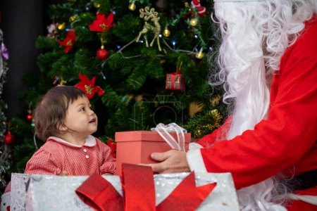 Foto de Bebé caucásico está recibiendo regalo de Santa Claus por la noche por el árbol de Navidad totalmente decorado para la celebración de la temporada - Imagen libre de derechos