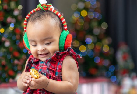 Foto de Bebé afroamericano es feliz sonriendo como jugar con adorno bauble mientras se viste con vestido de navidad y sombrero de santa con árbol de navidad en la parte posterior para la celebración de la temporada - Imagen libre de derechos