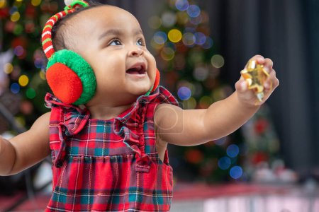 Téléchargez les photos : Afro-américain bébé est heureux de sourire comme jouer avec ornement boule tout en s'habillant en robe de Noël et santa chapeau avec arbre de Noël sur le dos pour la célébration de la saison - en image libre de droit