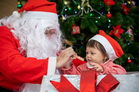 Téléchargez les photos : Le bébé caucasien reçoit un cadeau du Père Noël la nuit près de l'arbre de Noël entièrement décoré pour la célébration de la saison - en image libre de droit