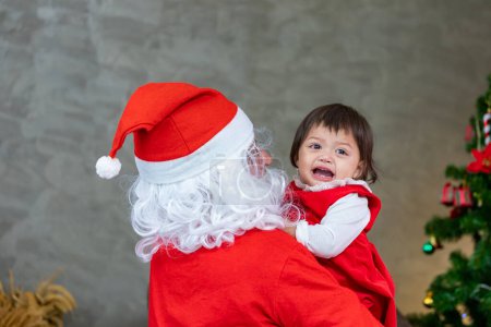 Téléchargez les photos : Père Noël soulève heureuse petite fille tout-petit bébé et rire gaiement avec arbre de Noël entièrement décoré sur le dos pour le concept de célébration de la saison - en image libre de droit