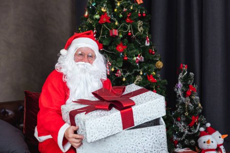 Foto de Santa Claus is holding christmas present box with fully decorated chrsitmas tree for season celebration and happy new year event - Imagen libre de derechos