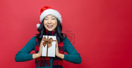 Téléchargez les photos : Jolie femme asiatique souriante en pull de Noël chaud et chapeau santa tenant boîte cadeau comme cadeau avec fond rouge pour le concept de célébration de la saison - en image libre de droit