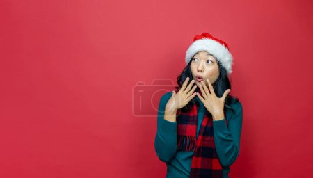 Foto de Bastante sonriente asiático mujer en caliente navidad suéter y santa sombrero es tener emoción en rojo fondo para temporada celebración concepto - Imagen libre de derechos