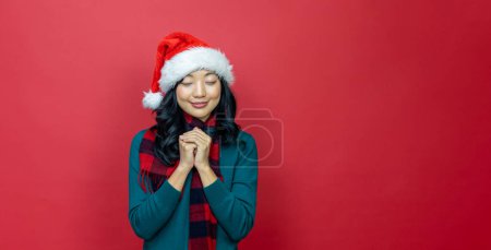 Téléchargez les photos : Jolie femme asiatique souriante en pull de Noël chaud et chapeau de Père Noël fait un souhait sur fond rouge pour la célébration de la saison - en image libre de droit