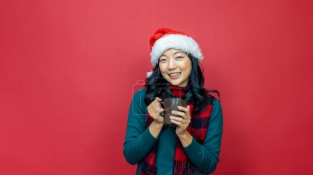 Téléchargez les photos : Jolie femme asiatique souriante en pull de Noël chaud et chapeau de Père Noël a chocolat chaud sur fond rouge pour la célébration de la saison - en image libre de droit