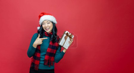 Téléchargez les photos : Jolie femme asiatique souriante en pull de Noël chaud et chapeau santa pointant vers la boîte cadeau comme présent avec fond rouge pour le concept de célébration de la saison - en image libre de droit